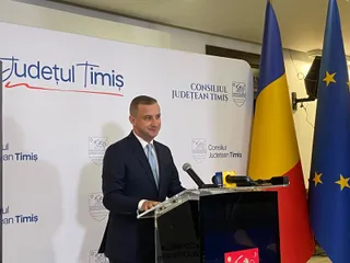 Un bărbat într-un costum închis la culoare și cravată deschisă stă în spatele unui podium cu microfoane, lângă steagurile României și Europei.
