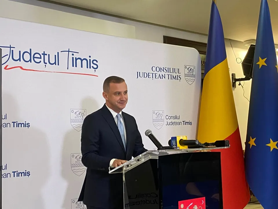 Un bărbat într-un costum închis la culoare și cravată deschisă stă în spatele unui podium cu microfoane, lângă steagurile României și Europei.