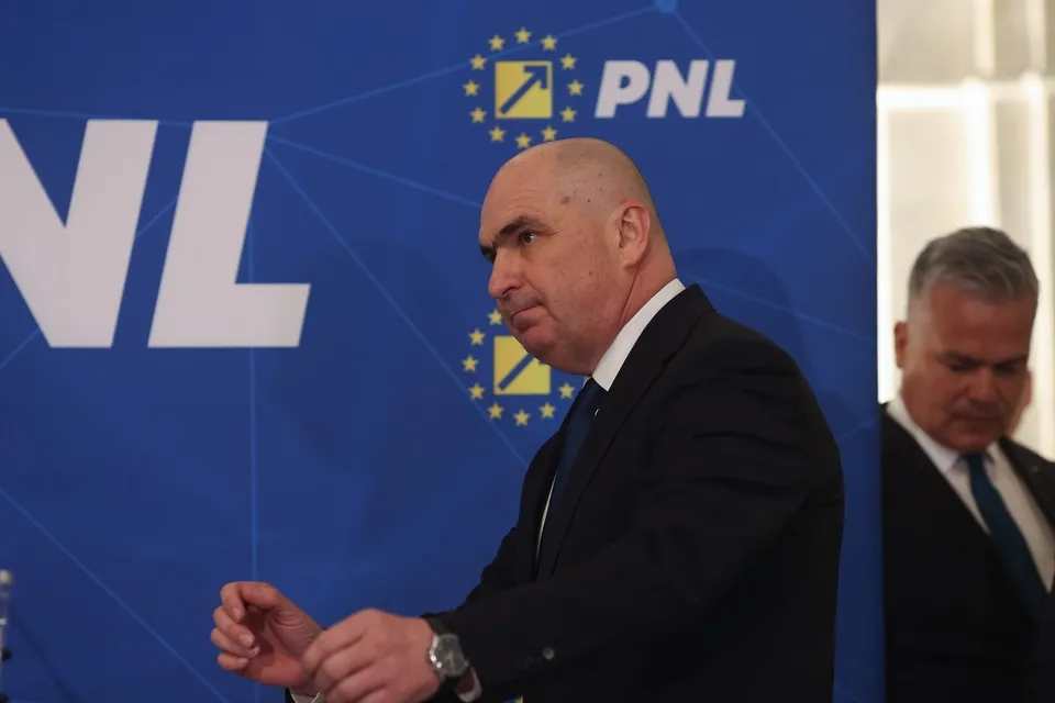 Un bărbat chel într-un costum negru și cămașă albă stă în fața unui panou albastru cu inscripții albe și galbene.