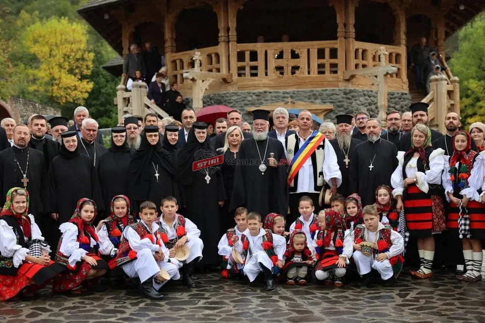 Un grup de oameni îmbrăcați în haine tradiționale și în robe negre stau grupați în fața unei construcții din lemn cu elemente sculptate.