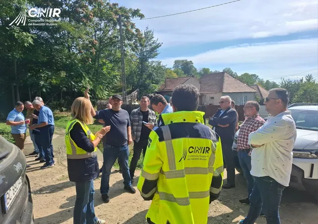 Un grup de persoane, inclusiv un bărbat cu vestă reflectorizantă CNIR, stau în aer liber, în apropierea unor case și copaci.