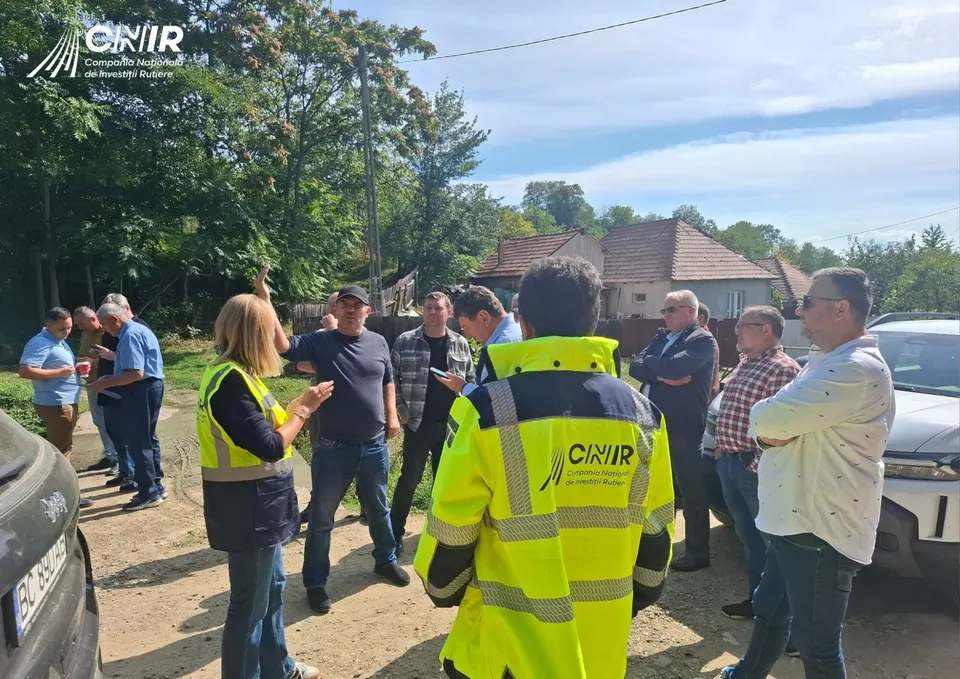 Un grup de persoane, inclusiv un bărbat cu vestă reflectorizantă CNIR, stau în aer liber, în apropierea unor case și copaci.