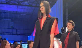 O femeie cu părul scurt poartă un vestă neagră peste o cămașă roșie transparentă, mergând pe o pistă de prezentare de modă.