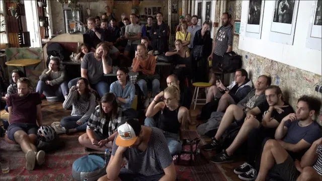 Un grup de persoane stă așezat pe podea și pe scaune într-o încăpere cu pereți decorați, privind spre o direcție comună.