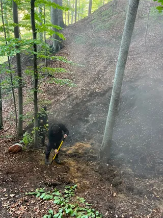 Un bărbat cu haine închise folosește un instrument cu mâner lung pe un teren înclinat, acoperit de pământ și vegetație într-o pădure.