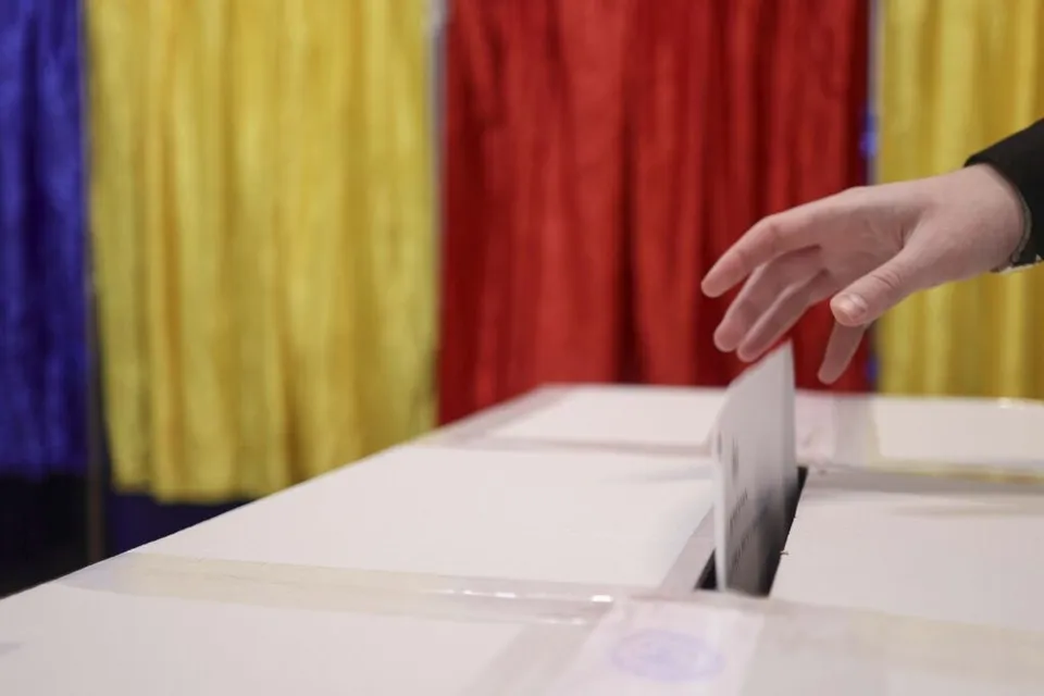 O mână introduce un plic alb într-o deschidere a unei cutii albe, în fața unor draperii colorate de culori galben, roșu și albastru.