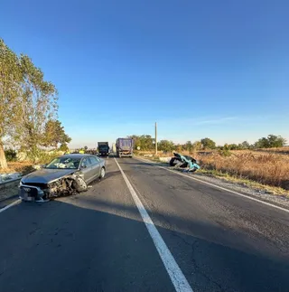 Un autoturism gri cu partea frontală distrusă stă pe un drum asfaltat lângă un alt vehicul de culoare închisă și resturi metalice.