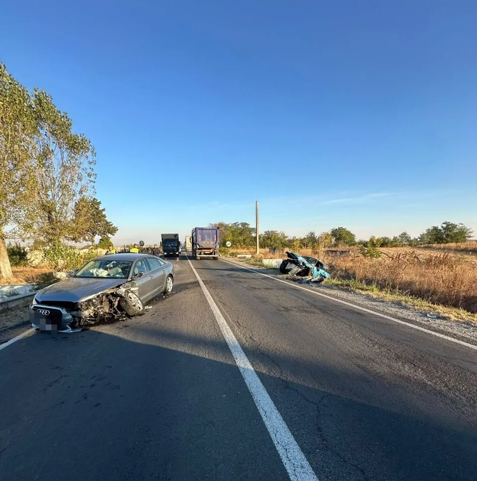 Un autoturism gri cu partea frontală distrusă stă pe un drum asfaltat lângă un alt vehicul de culoare închisă și resturi metalice.