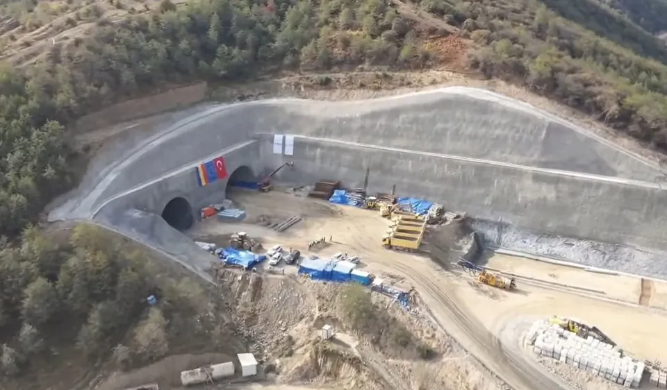O vedere aeriană prezintă două deschideri de tunel într-un versant de munte, cu utilaje de construcție și materiale de șantier dispuse pe un teren plat.