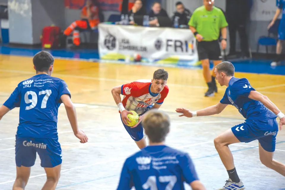 Un jucător de handbal în echipament roșu și alb conduce mingea pe terenul de joc, fiind urmărit de trei jucători în echipamente albastre.