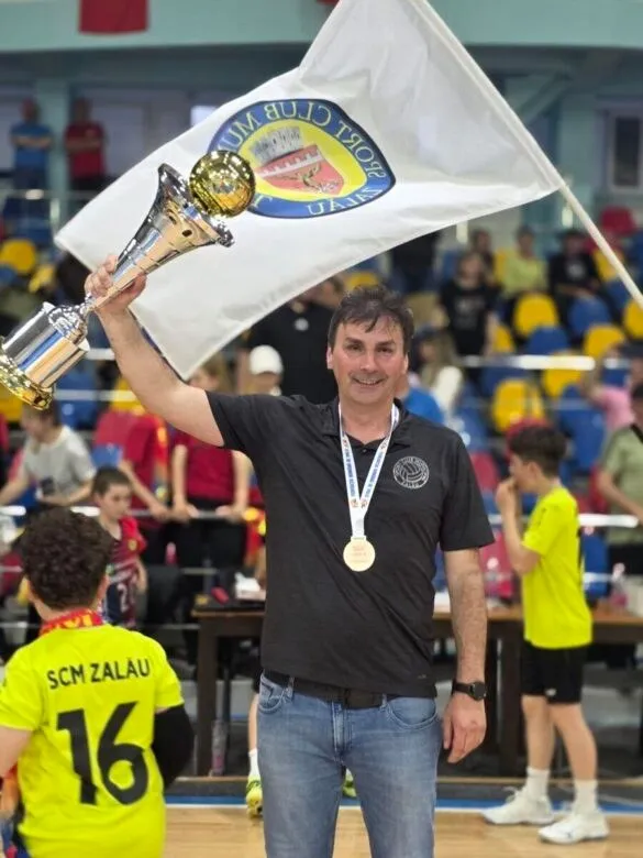 Un bărbat cu medalie aurie și tricou negru ridică un trofeu argintiu într-o sală de sport cu oameni în fundal.