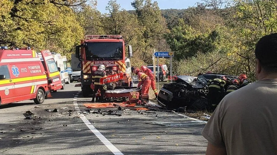 Pompieri în echipament de protecție și personal medical lucrează lângă un vehicul accidentat pe un drum asfaltat, în prezența unei ambulanțe și a unei autospeciale.