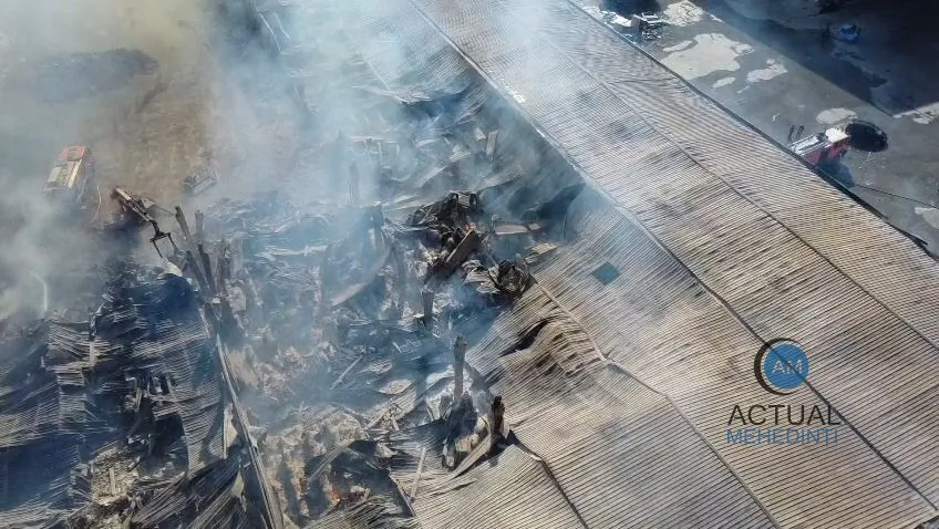 O vedere aeriană prezintă un clădire cu acoperiș metalic parțial distrus și zone acoperite de fum dens.