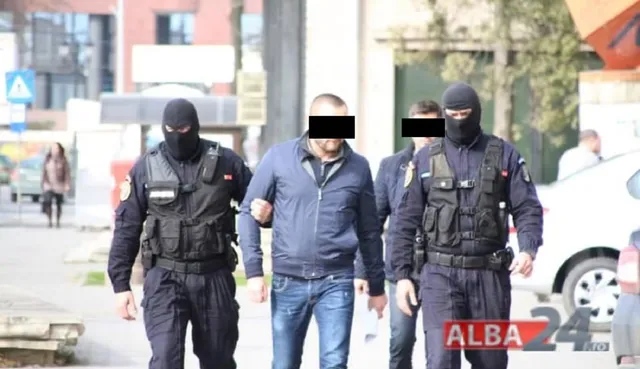 Trei bărbați în uniformă neagră cu măști și veste tactice conduc un bărbat cu sacou albastru și blugi pe un drum urban.