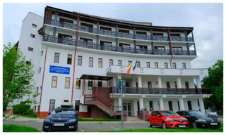 O clădire albă cu mai multe etaje, balcoane lungi și mașini parcate în fața acesteia sub un cer înnorat.