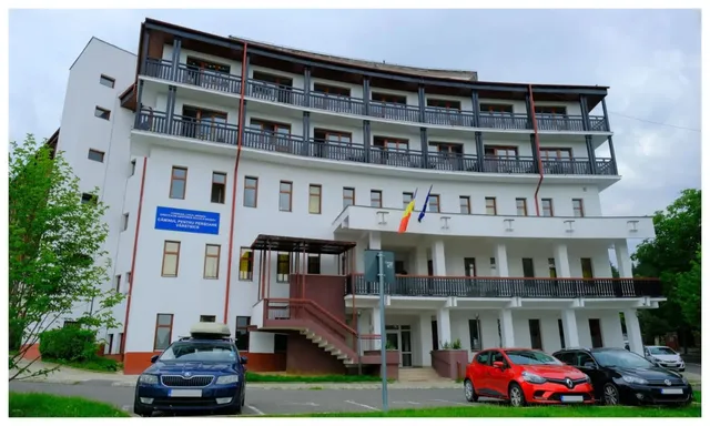 O clădire albă cu mai multe etaje, balcoane lungi și mașini parcate în fața acesteia sub un cer înnorat.