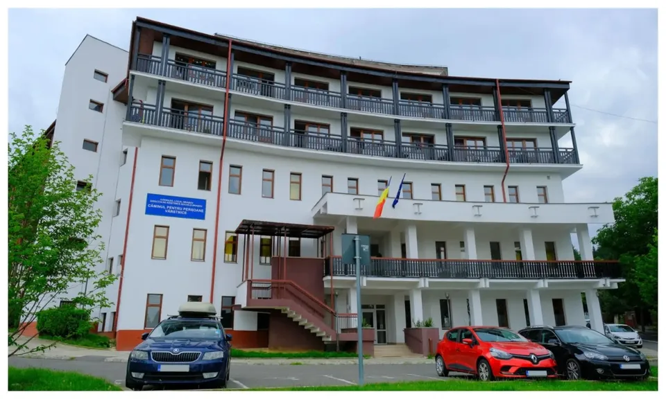 O clădire albă cu mai multe etaje, balcoane lungi și mașini parcate în fața acesteia sub un cer înnorat.