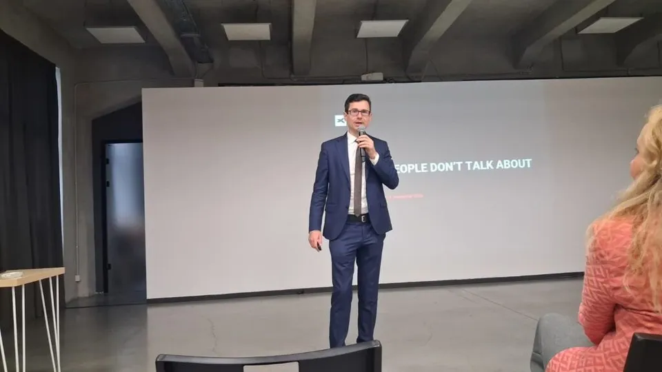Un bărbat cu ochelari și costum în bleu deschis stă în picioare pe o scenă, ținând un microfon în fața unui ecran alb mare.