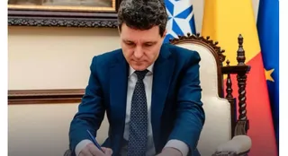 Un bărbat cu părul închis la culoare, purtând sacou albastru și cravată cu puncte, scrie pe o hârtie cu un pix.
