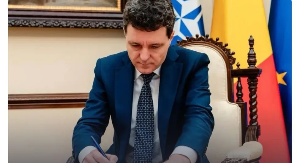 Un bărbat cu părul închis la culoare, purtând sacou albastru și cravată cu puncte, scrie pe o hârtie cu un pix.