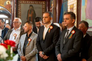 Câțiva bărbați în costume și cămașă stau în picioare într-o încăpere decorată cu picturi religioase, purtând rozete colorate pe piept.