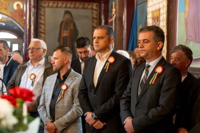 Câțiva bărbați în costume și cămașă stau în picioare într-o încăpere decorată cu picturi religioase, purtând rozete colorate pe piept.