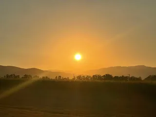 Soarele strălucește pe un cer portocaliu deasupra unor dealuri întunecate și a unei linii de copaci.