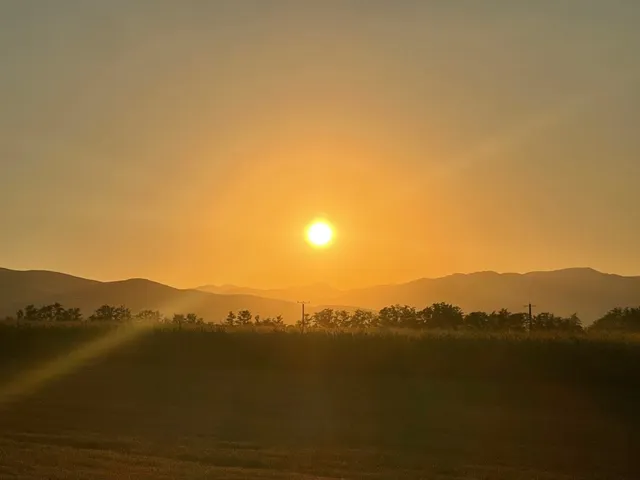 Soarele strălucește pe un cer portocaliu deasupra unor dealuri întunecate și a unei linii de copaci.