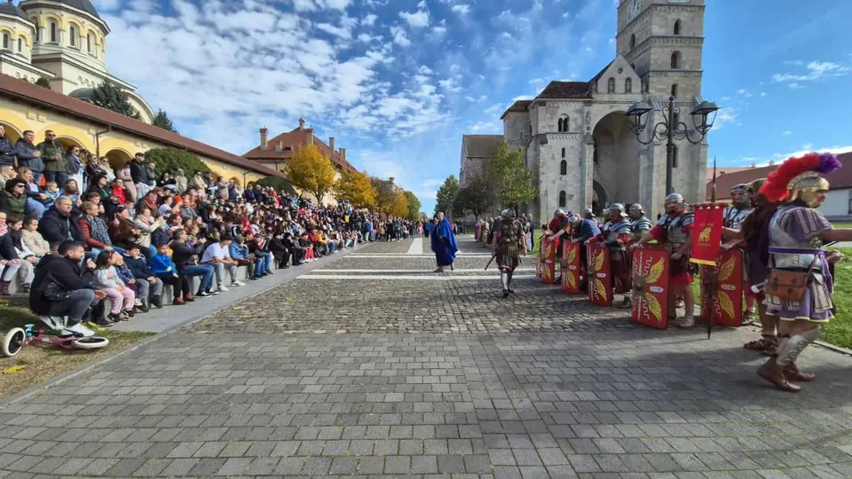 Oamenii stau așezați pe bănci în fața unei clădiri, privind spre un grup de persoane îmbrăcate în costume istorice pe o piață pavată.
