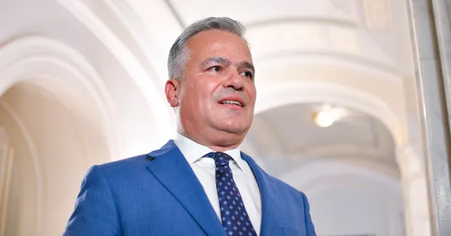Un bărbat cu părul cărunt, purtând sacou albastru, cămașă albă și cravată cu puncte, privește în sus într-un spațiu cu arcade albe.