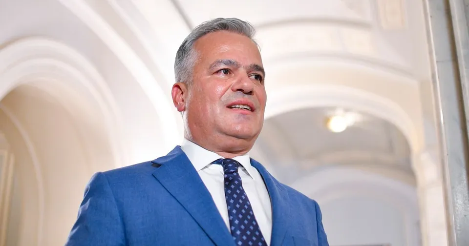 Un bărbat cu părul cărunt, purtând sacou albastru, cămașă albă și cravată cu puncte, privește în sus într-un spațiu cu arcade albe.