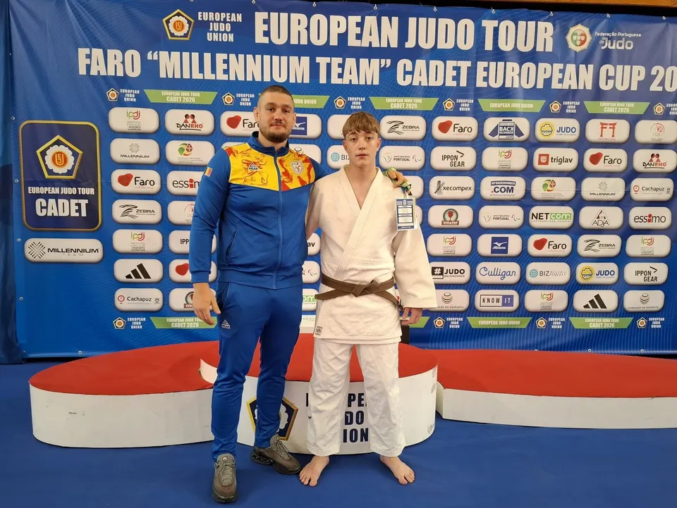 Un bărbat într-un costum sportiv albastru și galben stă lângă un tânăr îmbrăcat într-o kimonă albă în fața unui panou cu diverse logo-uri.