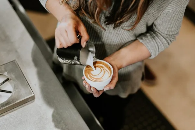 O persoană toarnă lapte dintr-un ibric metalic într-o cană albă de cafea pentru a crea un model pe spumă.