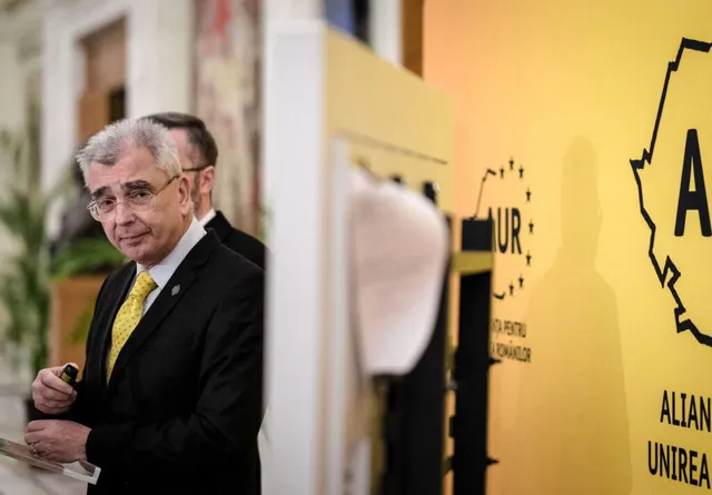 Un bărbat cu ochelari și costum negru cu cămașă albă și cravată galbenă stă lângă un panou galben cu text și simboluri negre.