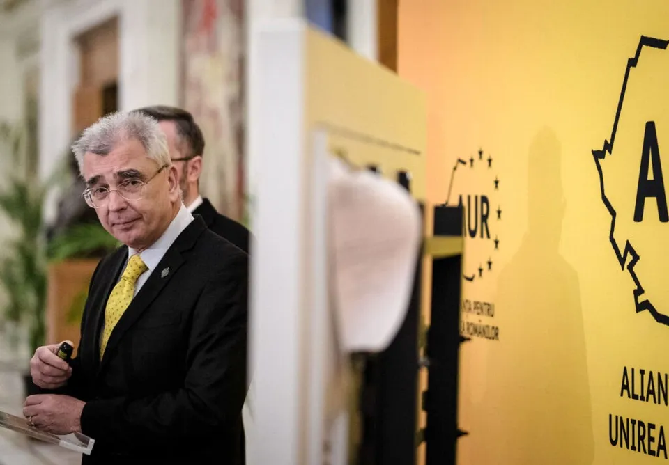 Un bărbat cu ochelari și costum negru cu cămașă albă și cravată galbenă stă lângă un panou galben cu text și simboluri negre.