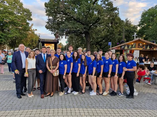 Un grup de persoane stă în rând pe o esplanadă pavată, în fața unor copaci și a unor construcții din lemn.