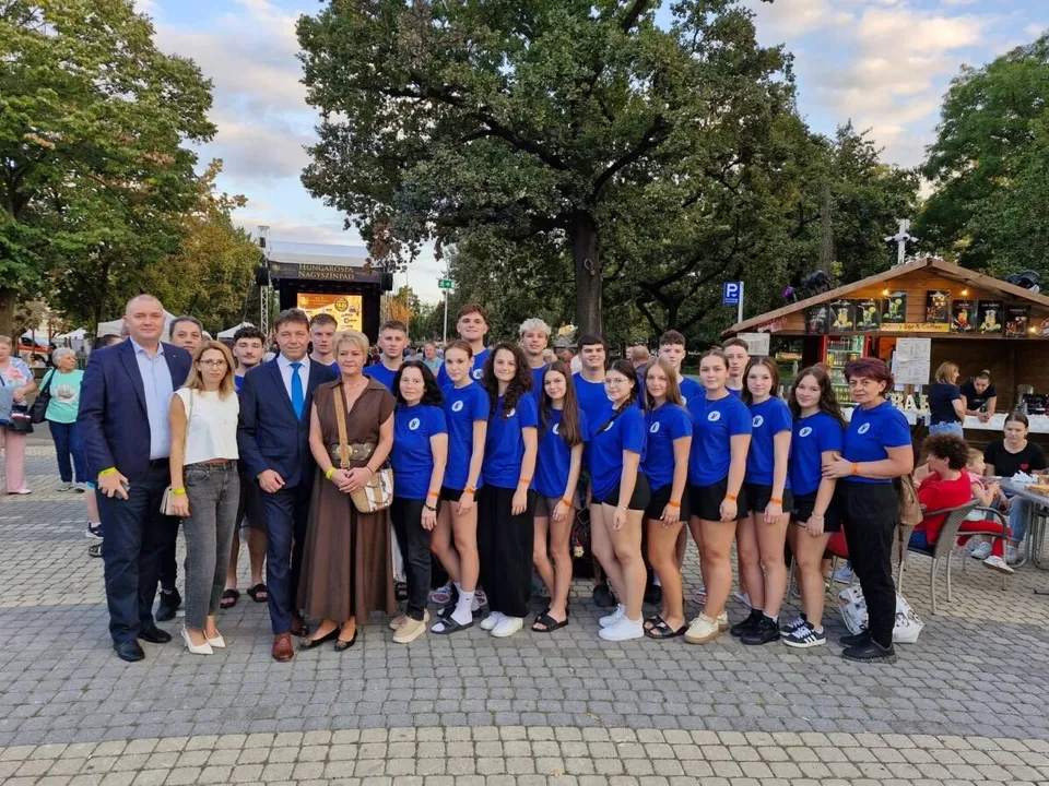 Un grup de persoane stă în rând pe o esplanadă pavată, în fața unor copaci și a unor construcții din lemn.