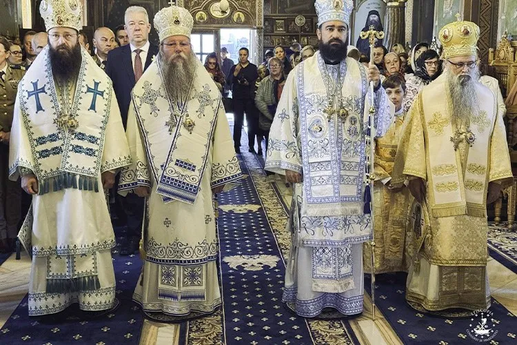Patru bărbați în haine liturgice albe cu broderii aurii și albastre stau în rând într-un interior decorat cu mochete albastre.