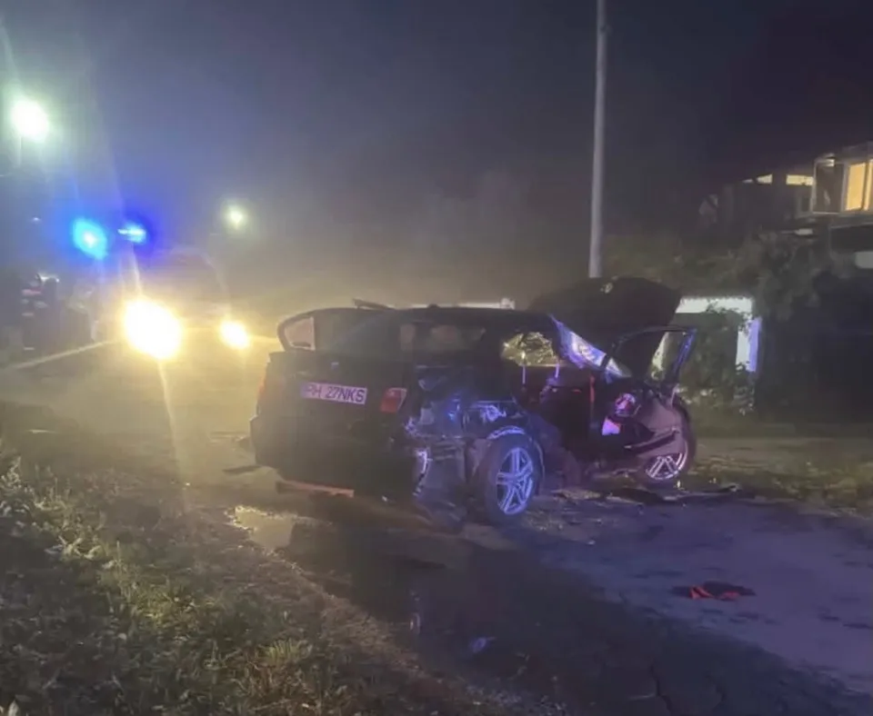 O mașină neagră, grav avariată, este parcată pe partea unei drumuri la noapte, cu capota și portbagaul deschise, în luminile intermitente ale unui vehicol de urgență.