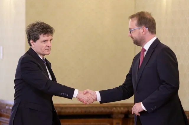 Doi bărbați în costume în plante și cravate se strâng în mână în fața unui perete de culoare deschisă.