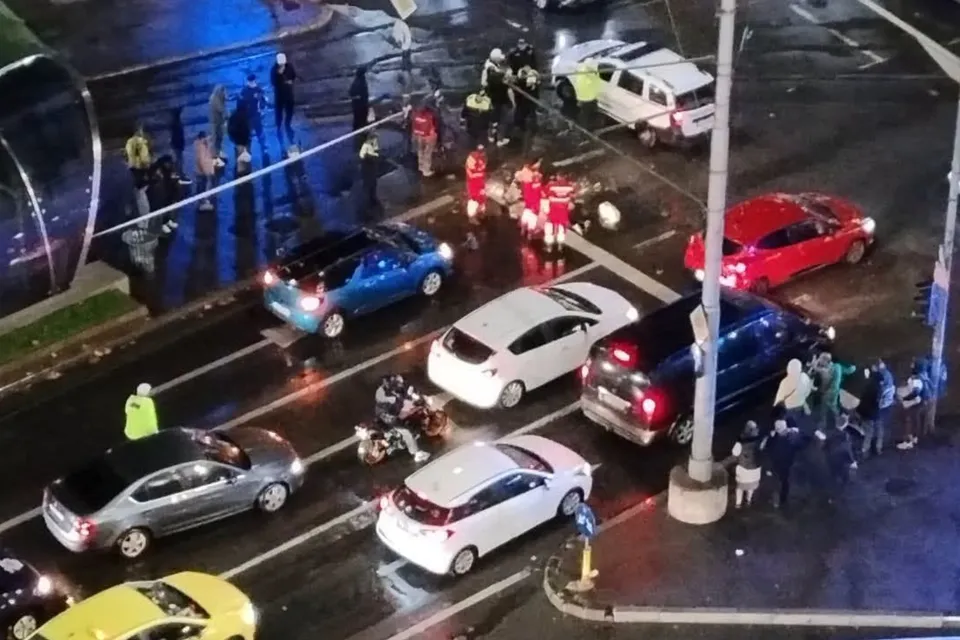 Imaginea de sus arată o intersecție aglomerată cu mai multe mașini, motociclete și persoane în uniformă, într-o scenă de accident rutier.