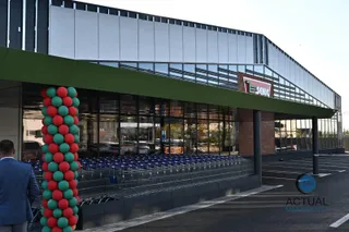 O clădire comercială modernă cu fațadă de sticlă este decorată cu un stâlp format din baloane roșii și verzi lângă un parcare asfaltată.