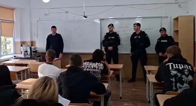 Patru bărbați în uniforme negri stau în fața unui grup de elevi așezați la bănci într-o sală de clasă cu tablă albă.