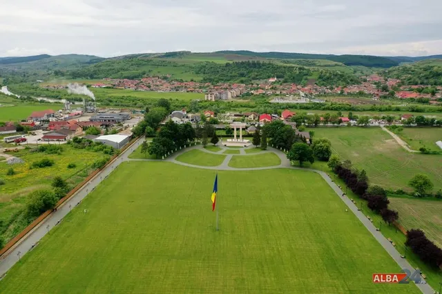 O vedere aeriană prezintă un câmp verde mare cu un stâlp de steag și un monument în depărtare, înconjurat de case și dealuri verzi.