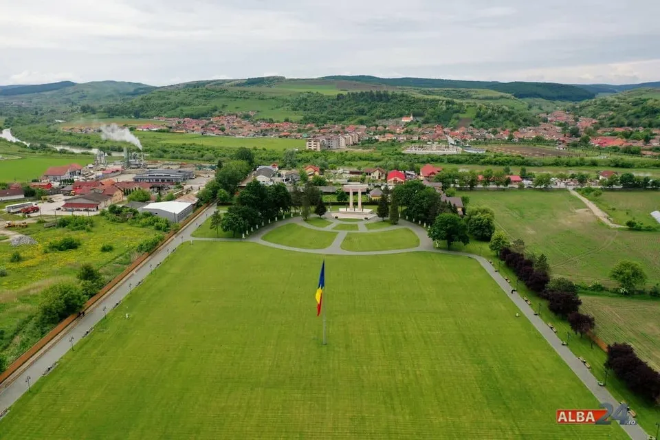 O vedere aeriană prezintă un câmp verde mare cu un stâlp de steag și un monument în depărtare, înconjurat de case și dealuri verzi.