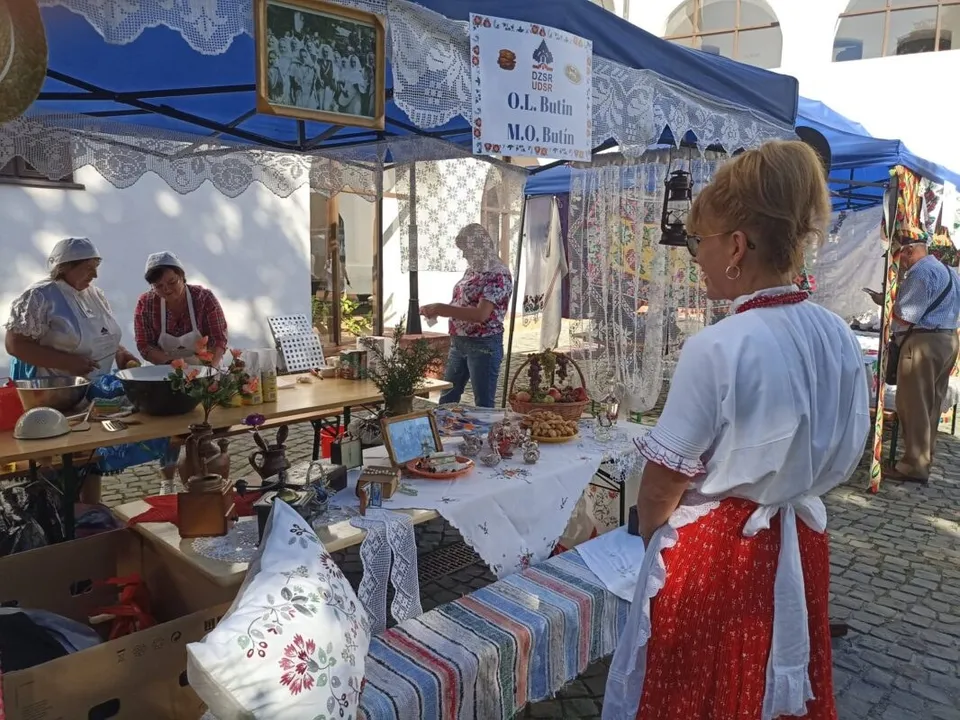 O femeie în costum tradițional roșu și alb stă lângă mese cu obiecte de decor și textile într-un stand acoperit.