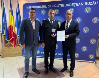 Trei bărbați stau în picioare în fața unui panou albastru cu embleme, unul purtând uniformă de poliție și ținând un obiect mic și o diplomă.