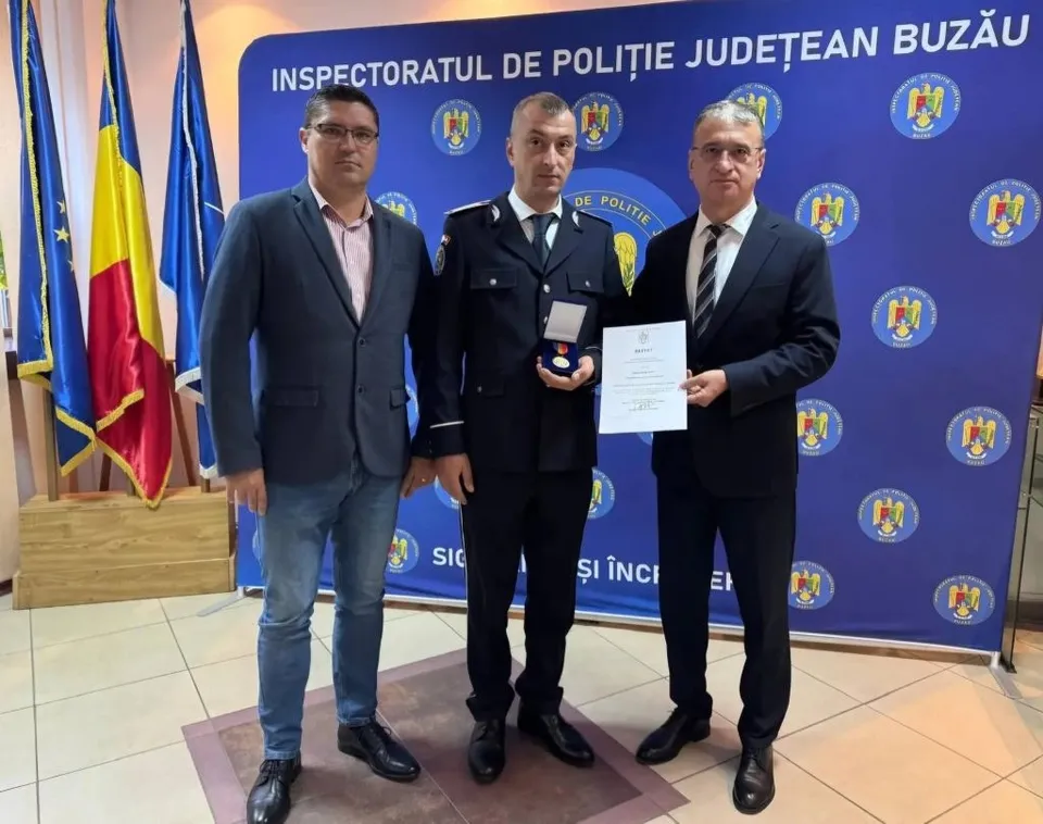Trei bărbați stau în picioare în fața unui panou albastru cu embleme, unul purtând uniformă de poliție și ținând un obiect mic și o diplomă.