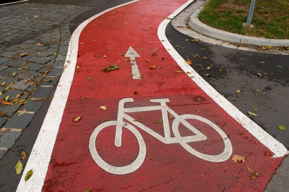 O pistă de biciclete roșie cu pictograme albe reprezentând o bicicletă și o săgeată este delimitată de linii albe pe un drum asfaltat.