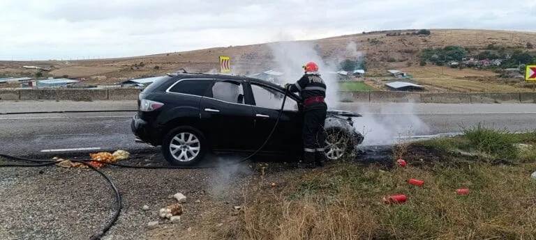 Un autoturism negru stă pe marginea unui drum lângă un teren cu iarbă, în timp ce un pompier în echipament de protecție stă lângă vehicul.
